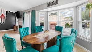 Un área de comedor con una mesa redonda, sillas tapizadas en color verde azulado, una chimenea y ventanas grandes con persianas.