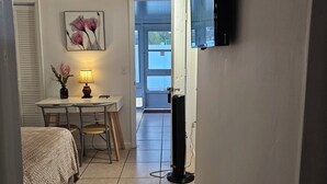 A small, well-lit room with a bed, a desk with a lamp, a TV mounted on the wall, and a door leading to an outdoor area. - Cozy studio, independent entrance, with AC in vibrant Orlando neighborhood (Orlando)