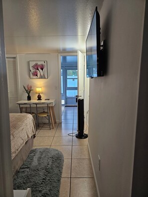 A small, well-lit room with a bed, a desk with a lamp, a TV mounted on the wall, and a door leading to an outdoor area. - Cozy studio, independent entrance, with AC in vibrant Orlando neighborhood (Orlando)