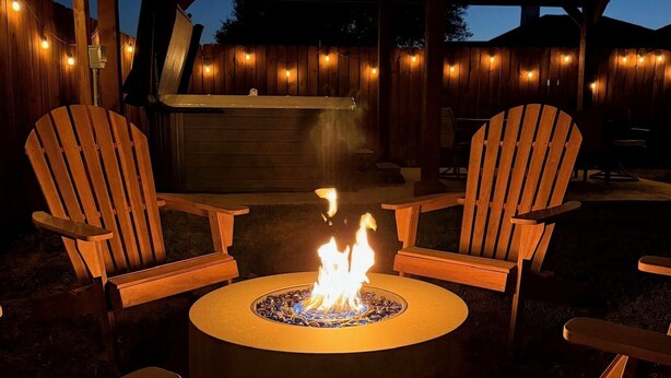 A circular fire pit with two wooden Adirondack chairs facing it, set against a wooden pergola with string lights. - The Olive Mesa - HotTub - Fire pit - Sandbox (Lubbock)