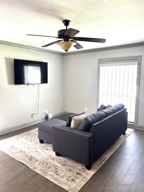 A living room with a flat-screen TV, a ceiling fan, a dark-colored sofa, and a patterned rug. - Cozy Graceful 2BR Near Downtown Houston (Houston)