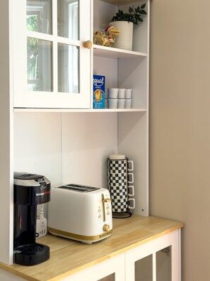 Una kitchenette con tostadora, cafetera, taza de cuadros y una planta en maceta sobre la repisa.