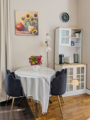 Et spiseområde med et rundt bord, to stole, en vase med blomster, et ur og et hvidt skab med hylder.
