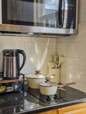 Una cocina con un hervidor de acero inoxidable, dos ollas con tapas de madera, un microondas y un recipiente para utensilios.
