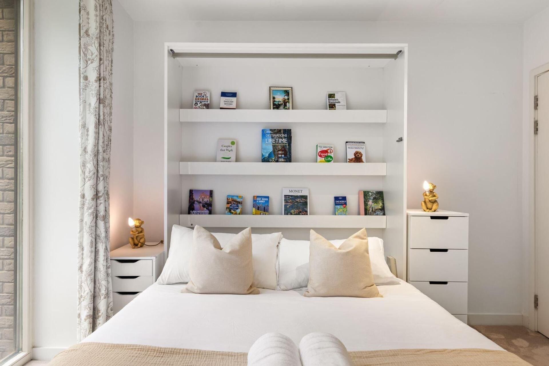 Een netjes opgeruimde slaapkamer met een bed, kussens, een nachtkastje, een boekenplank en een lamp.