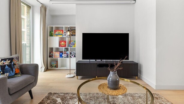 Ein modernes Wohnzimmer mit Flachbildfernseher, Bücherregal mit Gesellschaftsspielen, grauem Sofa und Couchtisch mit einer Vase.