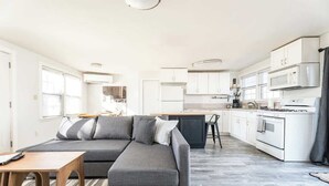 Une cuisine moderne avec des meubles blancs, un canapé gris et un coin repas doté d’une table en bois.