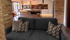 Uma sala de estar moderna com um sofá cinza escuro, almofadas estampadas e vista para uma cozinha com ilha central.
