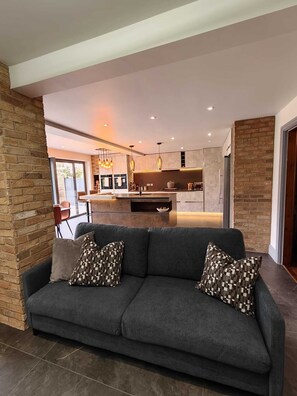 Ein modernes Wohnzimmer mit einem dunkelgrauen Sofa, gemusterten Kissen und Blick in einen Küchenbereich mit Kochinsel.