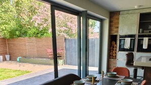 Uma área de jantar moderna com uma mesa comprida posta para seis pessoas, grandes janelas e vista para um quintal com cerca de madeira e vegetação.