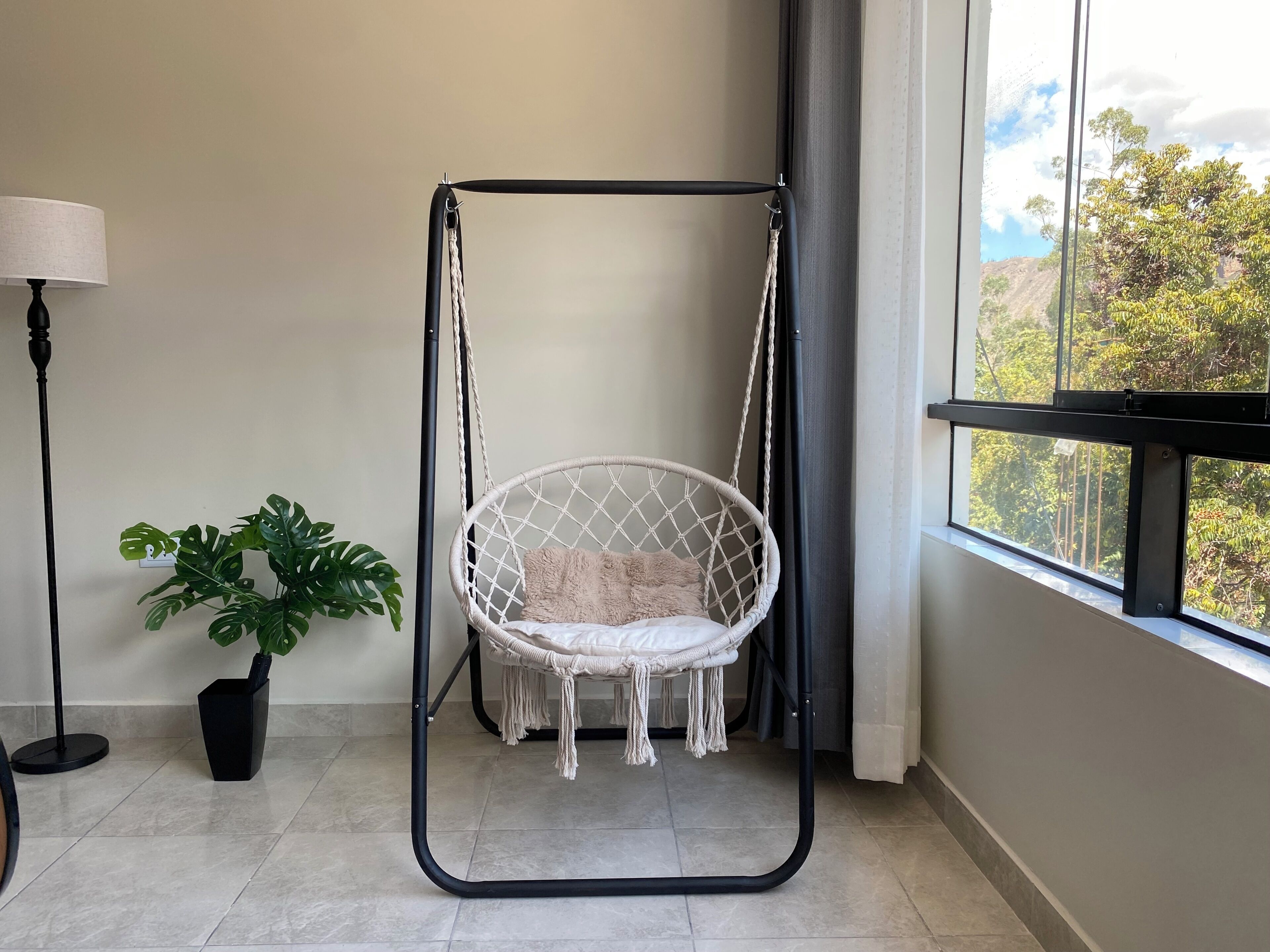 A room with a macrame hanging chair, a potted plant, a floor lamp, and a window with a view of trees.
