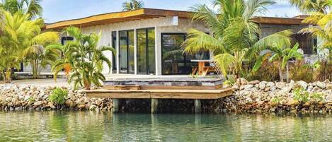 A modern house with a wooden deck over water, surrounded by palm trees.