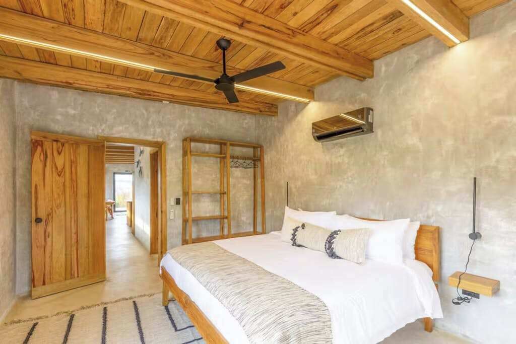 A bedroom with a wooden ceiling, a bed with white bedding, a ceiling fan, and a wall-mounted air conditioning unit.