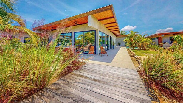 Un bâtiment moderne doté de grandes fenêtres, d’une terrasse en bois et de plantes tropicales.