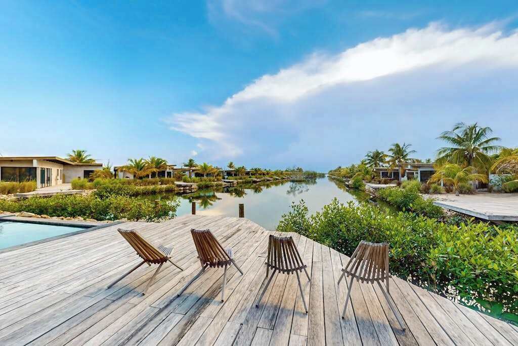 Een houten terras met lounge stoelen met uitzicht op een kalm wateroppervlak, omgeven door groen en moderne gebouwen.