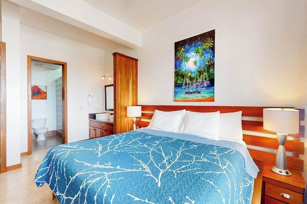 Habitación de hotel con una cama, un cuadro de una playa, un cabecero de madera y una mesita de noche con una lámpara.