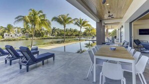 Ett modernt utomhusområde med plats för måltider, en pool, solstolar och utsikt över palmer och vatten.