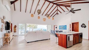 Un vaste espace de vie comprenant un grand canapé, des poutres apparentes, un îlot de cuisine et un ventilateur de plafond.