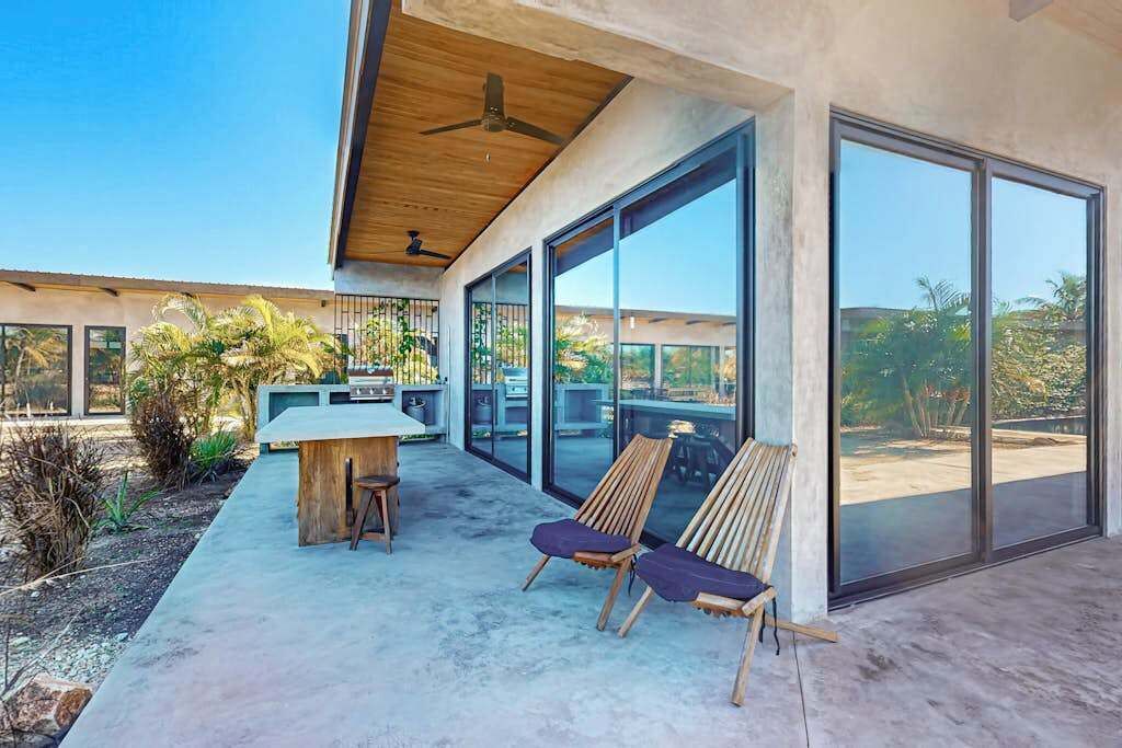 Una moderna terrazza esterna con tavolo e sedie in legno, ampie porte scorrevoli in vetro e una ventola a soffitto.