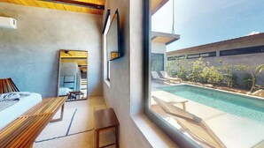 A modern hotel room with a large window overlooking a swimming pool, a bed, a desk, and a chair. - Elegant waterfront villa with private terrace, pool, beach access & 4 en-suites (Placencia)
