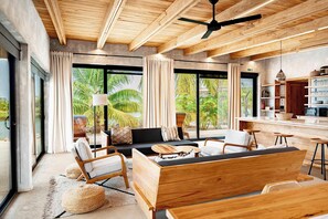 A modern living area with a wooden ceiling, large windows, a sofa, chairs, a coffee table, and a kitchen area with shelves. - Modern Waterfront Villa | Private Pool & Dock (Placencia)