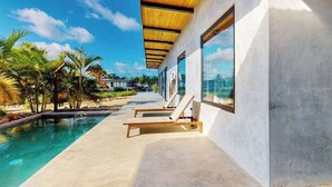A modern poolside area with lounge chairs, a concrete pool, and a view of palm trees and a clear sky. - Elegant waterfront villa with private pool, terrace and beach club access (Placencia)
