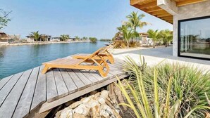 A wooden deck with a lounge chair, a modern building with a large window, and a body of water in the background. - Tranquil waterfront villa with private deck & full kitchen (Placencia)
