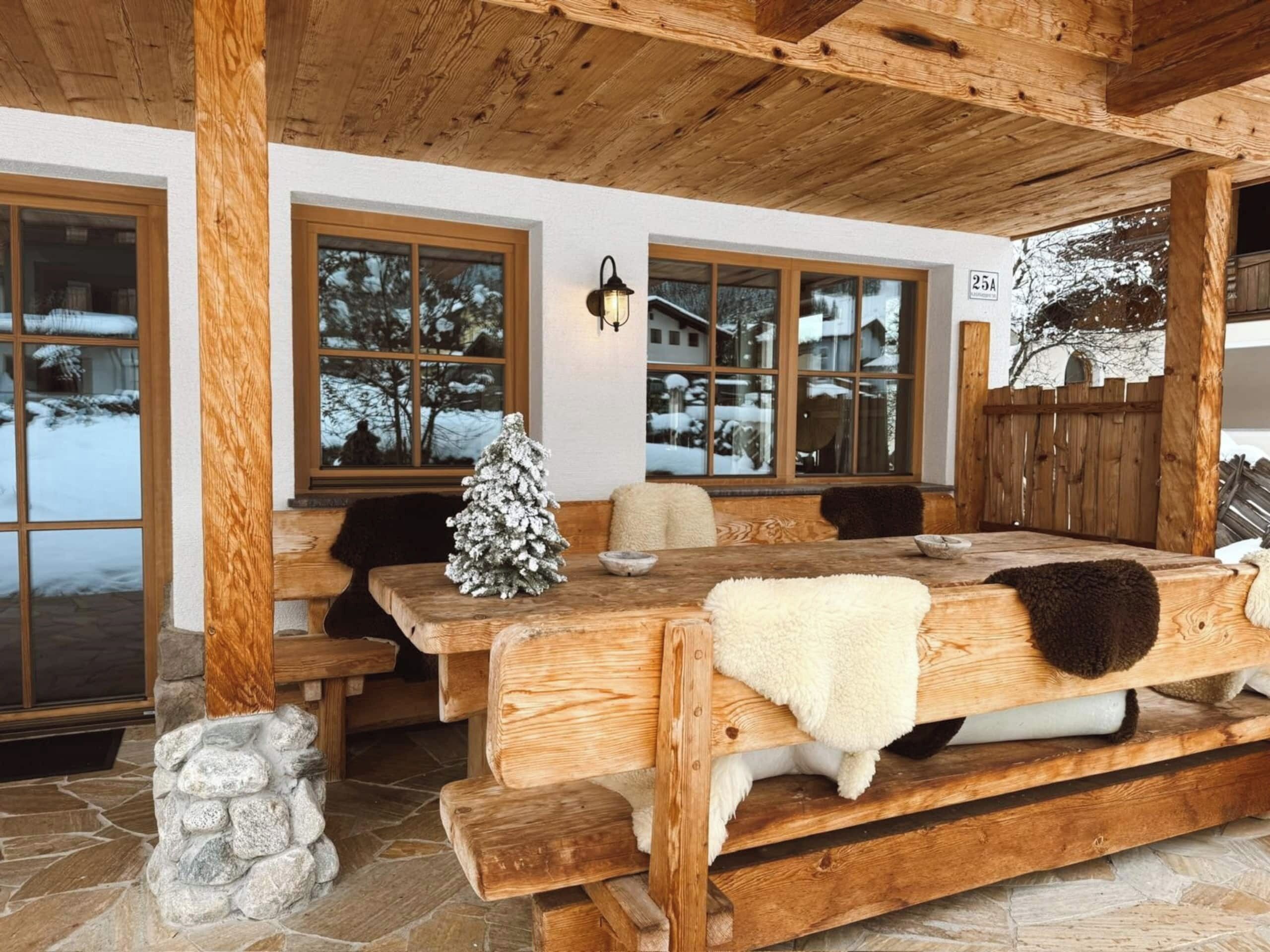 Eine hölzerne Bank mit einem weißen Fellüberwurf, ein Steinkamin und ein mit Schnee bedeckter Baum vor dem Fenster.