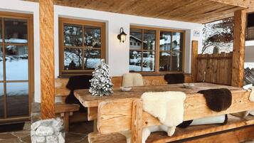 Eine hölzerne Bank mit einem weißen Fellüberwurf, ein Steinkamin und ein mit Schnee bedeckter Baum vor dem Fenster.