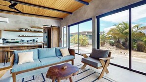 Ein modernes Interieur mit Sofa, Sessel, Couchtisch, großen Fenstern und einer Küchenzeile.
