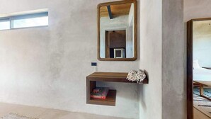 Ein Zimmer mit einer Betongwand, einem Holzbodenregal mit Büchern, einem Spiegel und einem Fenster.