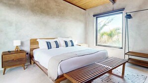Ein Schlafzimmer mit einem Bett, Nachttischen, einer Bank, einem Deckenventilator und einem großen Fenster mit Blick auf Palmen.