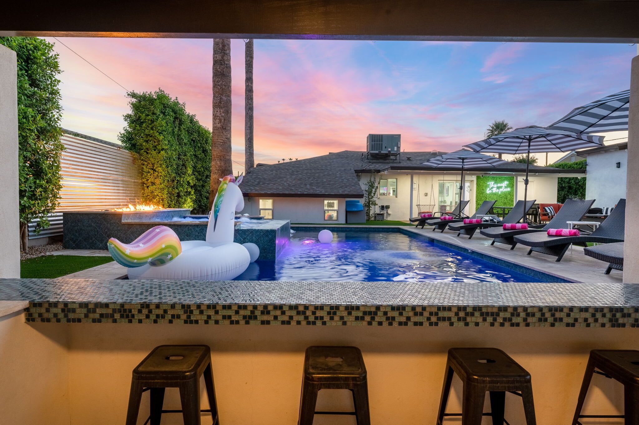 Un espace piscine avec des chaises longues, un flamant rose décoratif et un bar avec des tabourets.