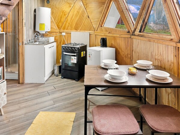 A compact kitchen with a stove, refrigerator, and a water heater. There is a dining table with plates and glasses, and a window with a view of the outdoors.