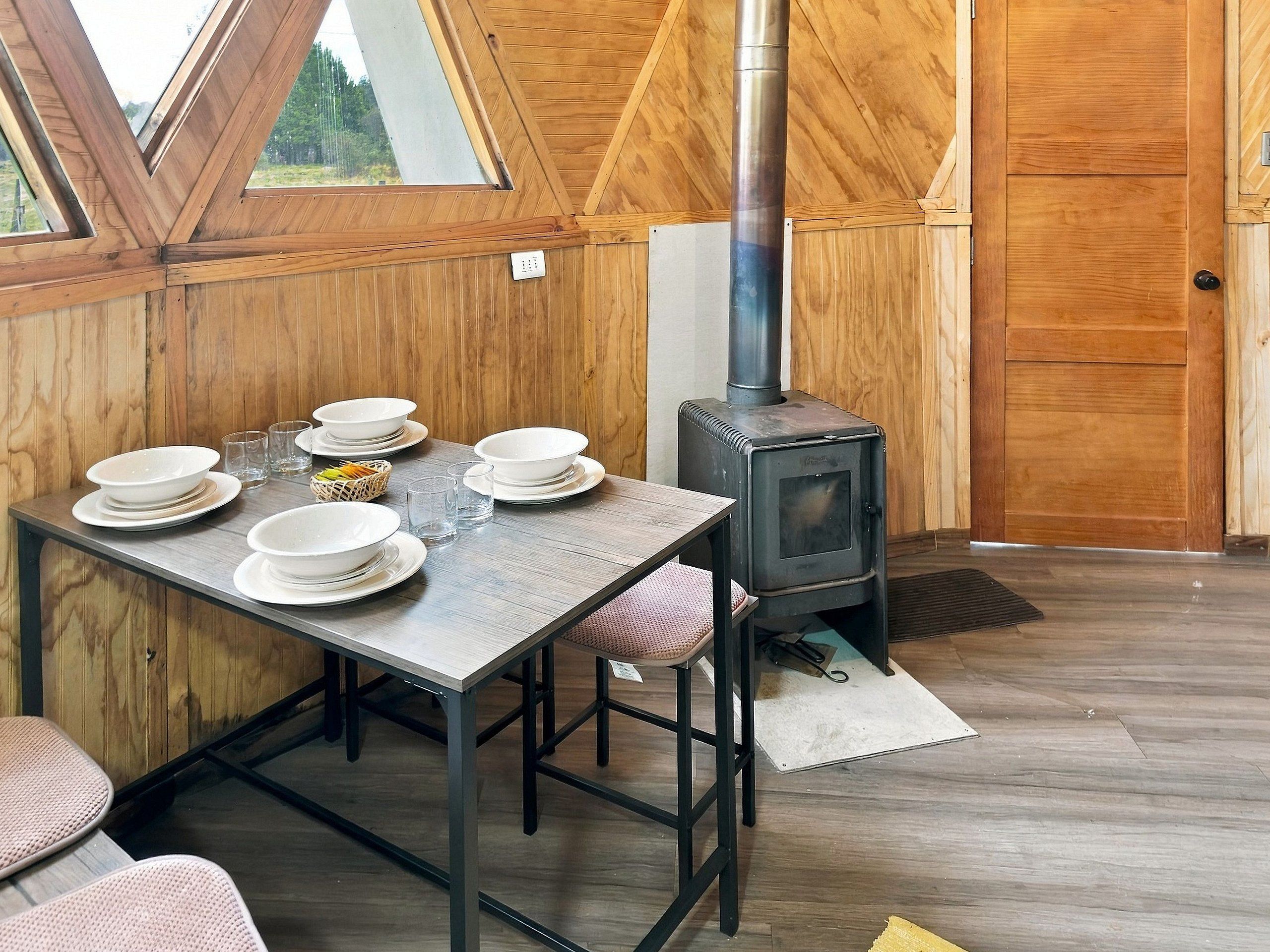 Un coin repas avec une table dressée pour quatre personnes, un poêle à bois et une vue à travers une fenêtre triangulaire.