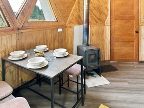 Un comedor con una mesa preparada para cuatro, un estufa de leña y vistas a través de un ventanal triangular.