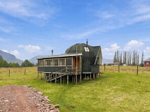 A small, single-story wooden structure with a sloped roof, situated in a grassy field with mountains in the background.