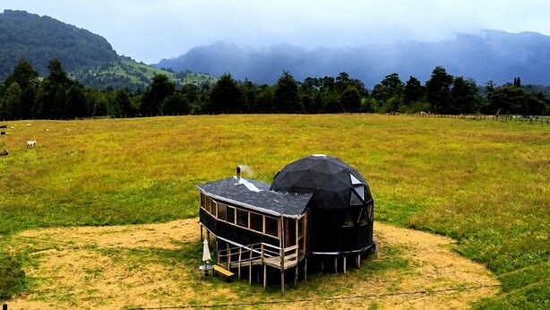 A geodesic dome structure in a grassy field with a wooden building nearby.
