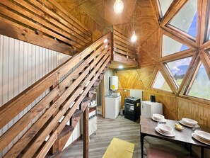 Un intérieur en bois avec un escalier, une table à manger dressée avec des assiettes, et une vue à travers une grande fenêtre.