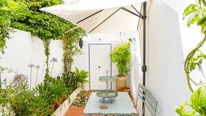 Eine kleine Terrasse im Freien mit Tisch und Stühlen, umgeben von Topfpflanzen und einer weißen Wand.