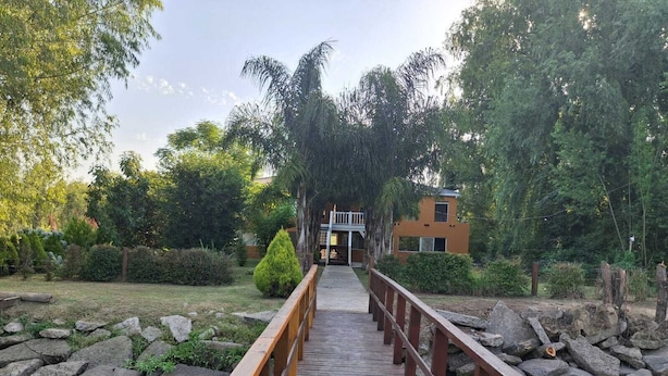 Un puente de madera que lleva a un edificio con balcón, rodeado de follaje y rocas.