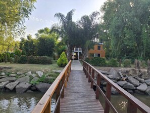 Eine Holzbrücke führt zu einem Gebäude mit einem Balkon, umgeben von Grünpflanzen und Felsen.
