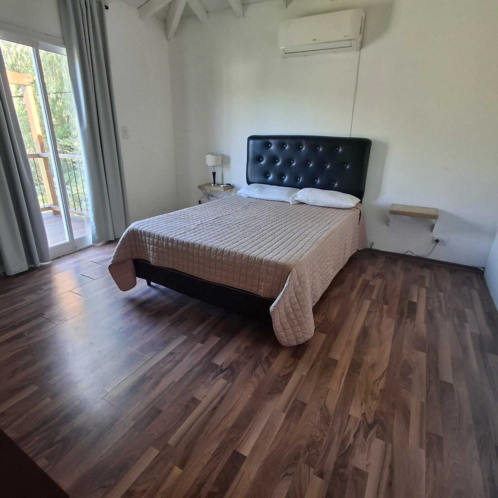 Un dormitorio con cama, suelo de madera, ventana con cortinas y aire acondicionado.