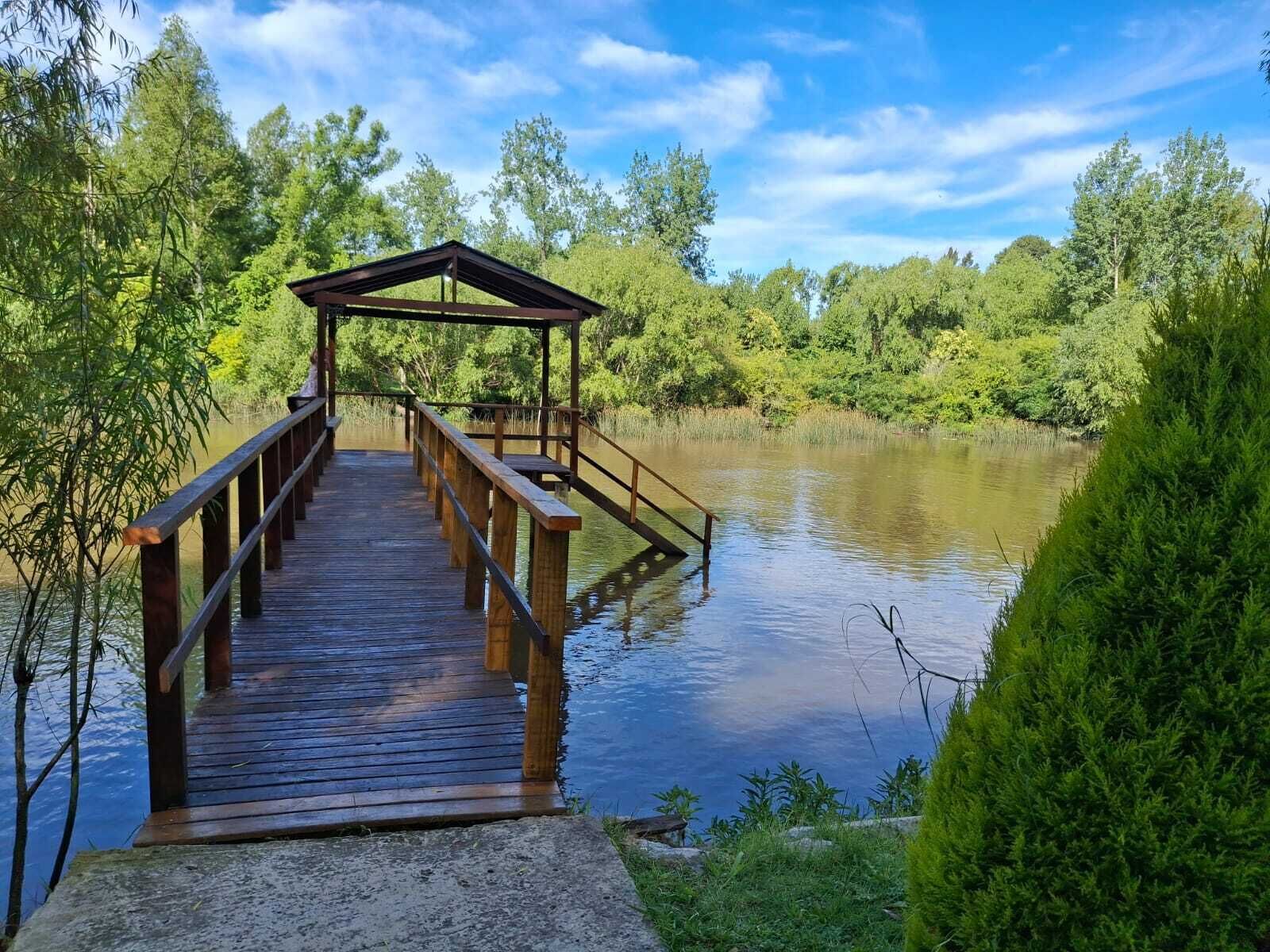 A wooden dock leading to a covered pavilion by a calm lake surrounded by lush greenery.