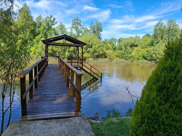 Un muelle de madera conduce a un pabellón cubierto junto a un lago tranquilo, rodeado de abundante vegetación.