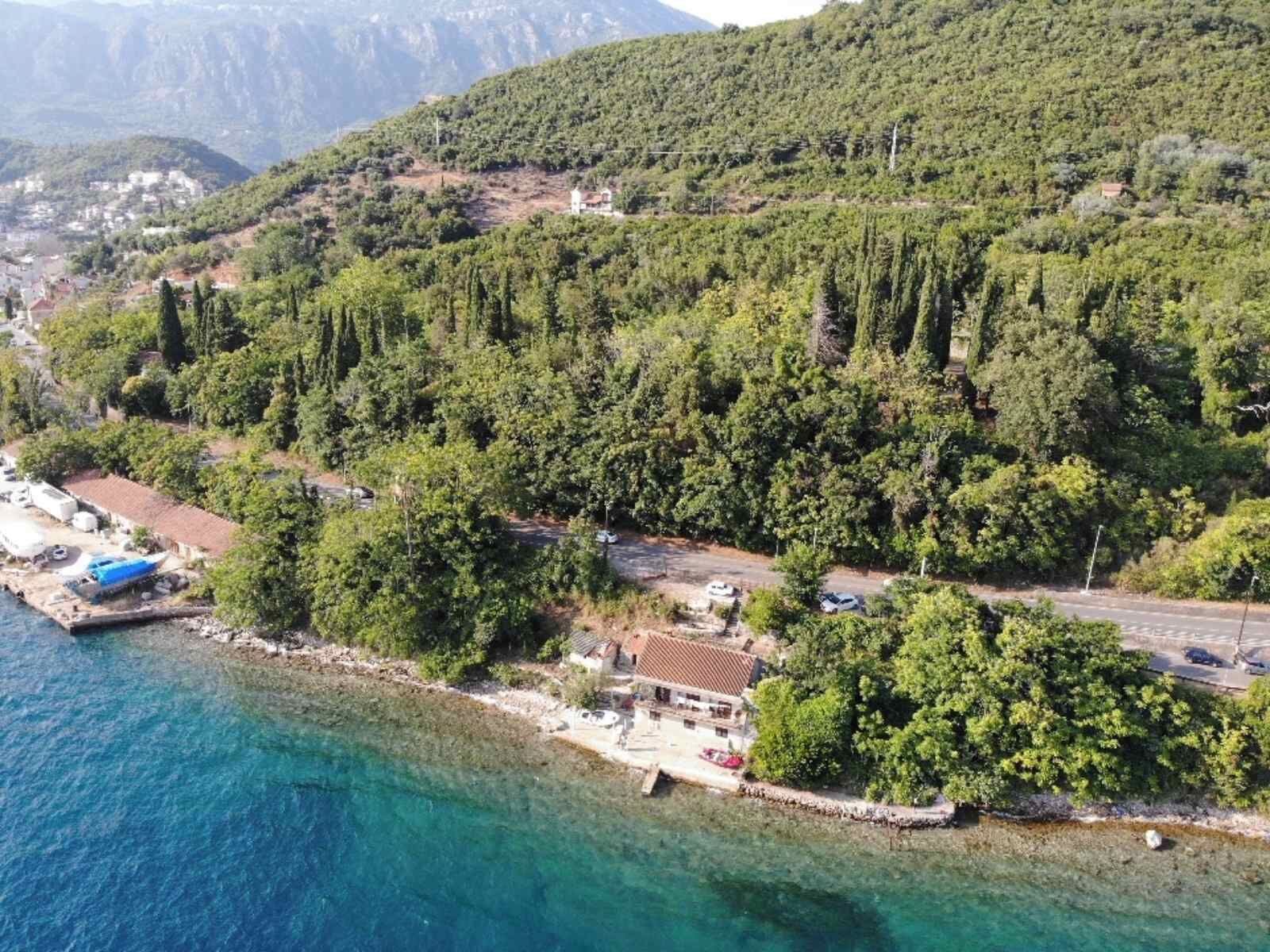 An aerial view of a coastal area with a small building near the water, surrounded by dense greenery and mountains in the background.