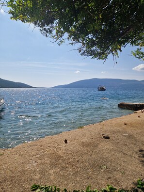 A beach with clear water, a boat, and distant mountains. - Charming 2-bedroom house with AC in peaceful Kumbor with private besch and boat (Kumbor)
