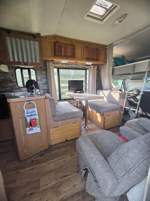A compact RV interior with a kitchenette, a small dining area, and seating.