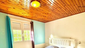 Ein Schlafzimmer mit Holzbalkendecke, einem Bett mit weißem Kopfteil, einem Fenster mit Vorhängen und einem Nachttisch mit Lampe.