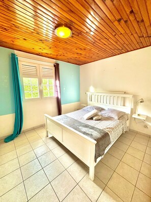 A bedroom with a wooden ceiling, a bed with a white headboard, a window with curtains, and a bedside table with a lamp. - Villa Solana, private pool with tropical garden in Sainte-Luce (Sainte-Luce)